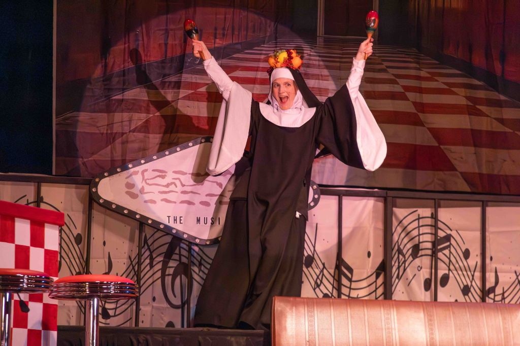 A performer dressed as a nun energetically poses on stage, holding maracas and wearing a colorful fruit headpiece, with a backdrop featuring musical notes.
