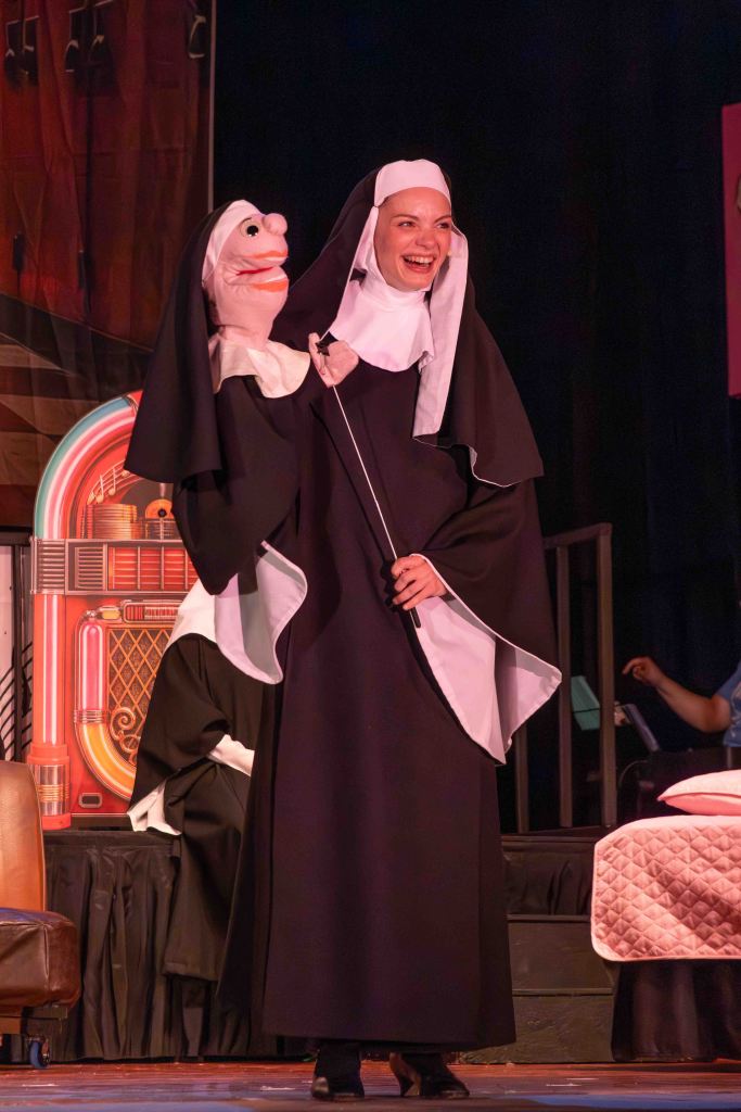 A performer dressed as a nun holds a puppet while smiling on stage during a theatrical performance.
