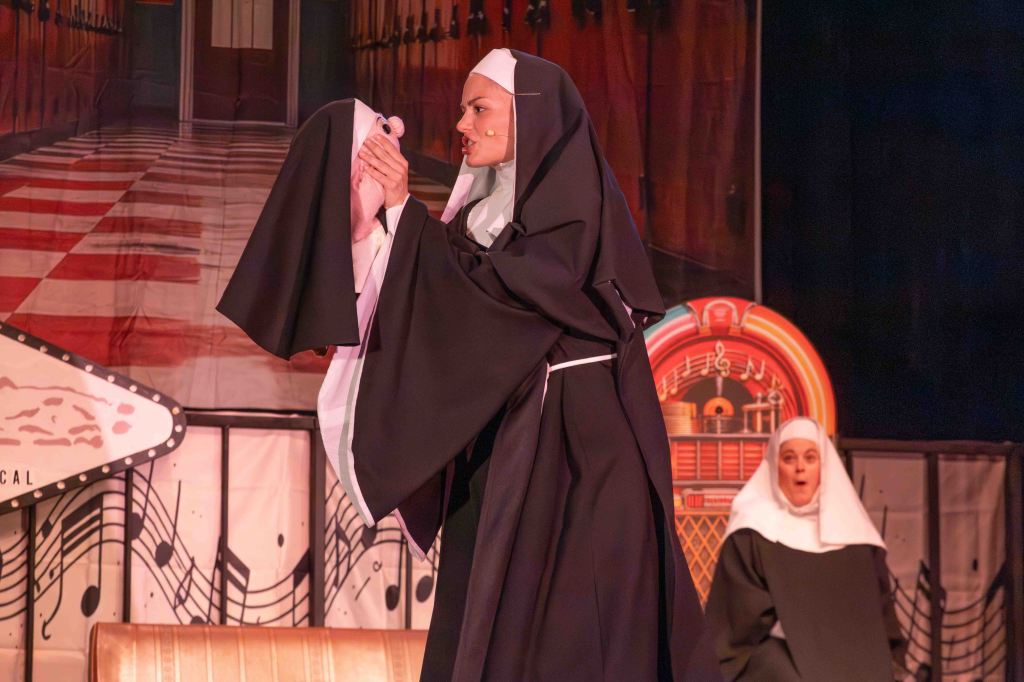 A woman dressed as a nun holds a stuffed animal while performing on stage, with another nun character looking surprised in the background. The stage backdrop features a hallway and musical elements.