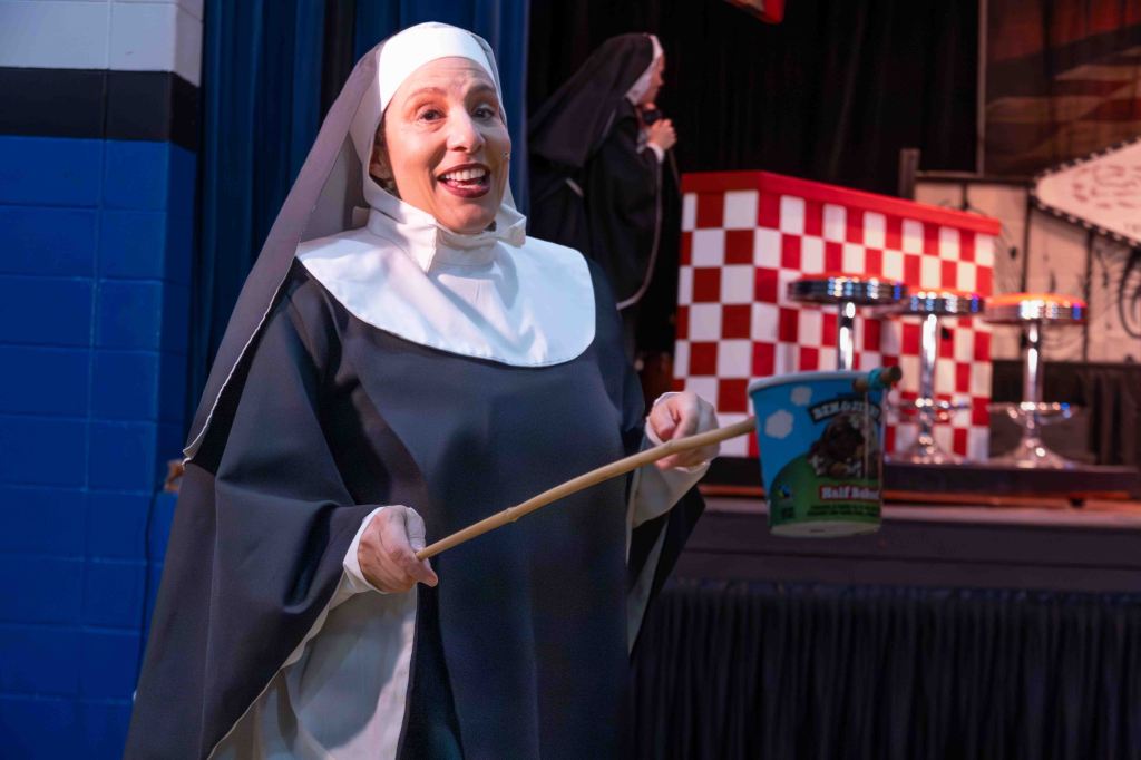 A smiling nun in traditional attire holding a stick and a colorful bucket, standing on stage with a checkered backdrop and bar stools.