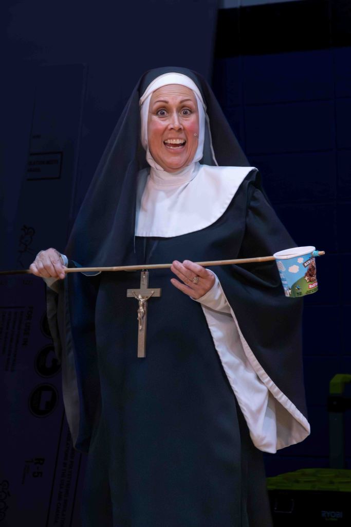A joyful nun wearing traditional habit holding a wooden stick with a cup attached, smiling and engaging with the audience.