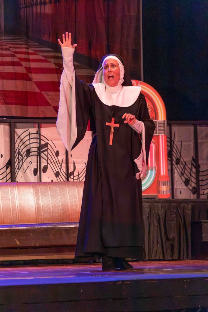 A nun in a black habit and white wimple raises her hand dramatically while performing on stage, with an expressive facial expression. The background features musical notes and a jukebox, indicating a lively theatrical atmosphere.
