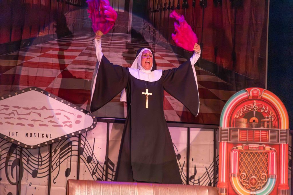 A joyful nun in a black habit raises her arms, holding pink feather boas, on stage with a vibrant backdrop and a jukebox prop.