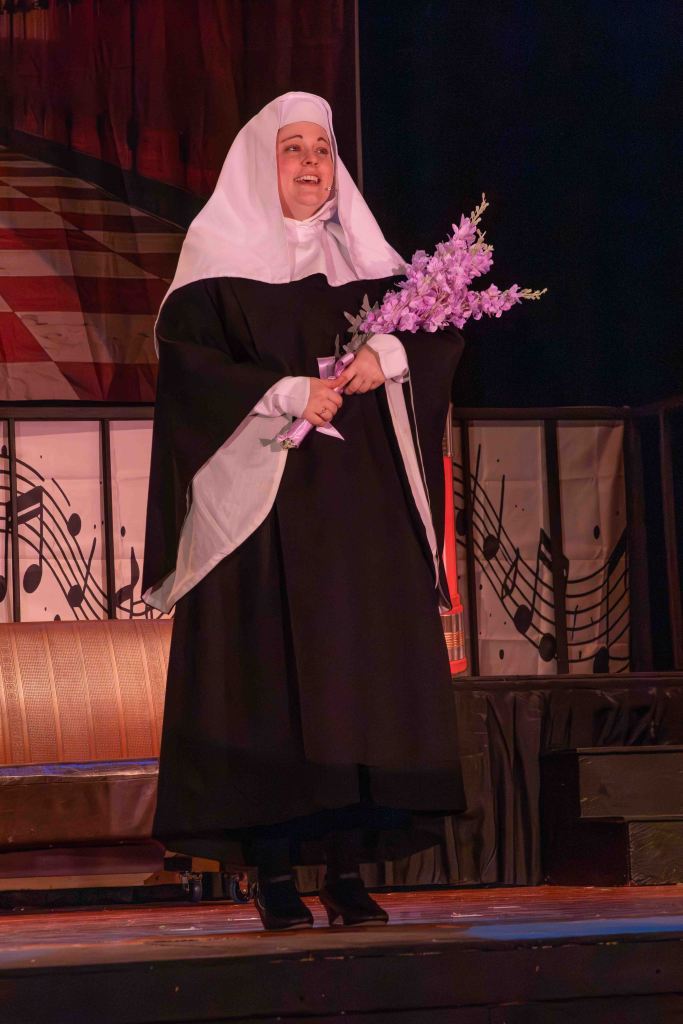 A woman dressed as a nun in a black habit and white veil, holding a bouquet of purple flowers, standing on stage with a joyful expression.