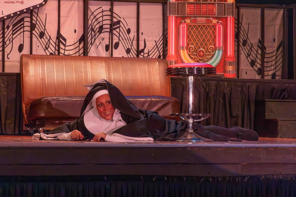 A performer in a nun costume lying on a stage, smiling playfully, with a brown couch and a jukebox backdrop.