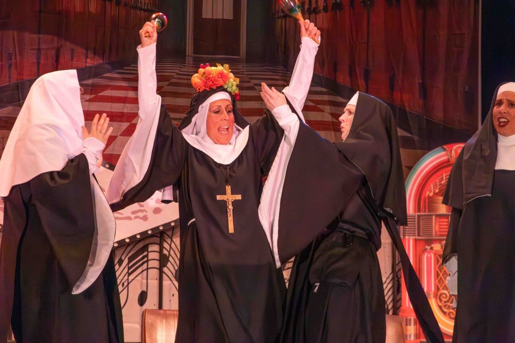 A lively theatrical scene featuring multiple nuns in traditional attire, one nun is joyfully holding a tambourine and wearing a floral crown while others participate in the performance.