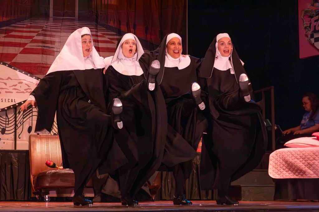 Four women dressed as nuns performing a lively dance on stage, showcasing energetic expressions and synchronized leg movements.