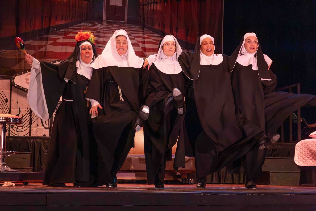 Five women dressed as nuns performing on stage, joyfully lifting their legs, with one holding a maraca, set against a colorful backdrop.