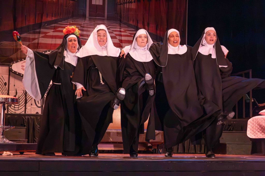 Five women in nun costumes dancing on stage with joyful expressions, holding props and performing a lively routine.