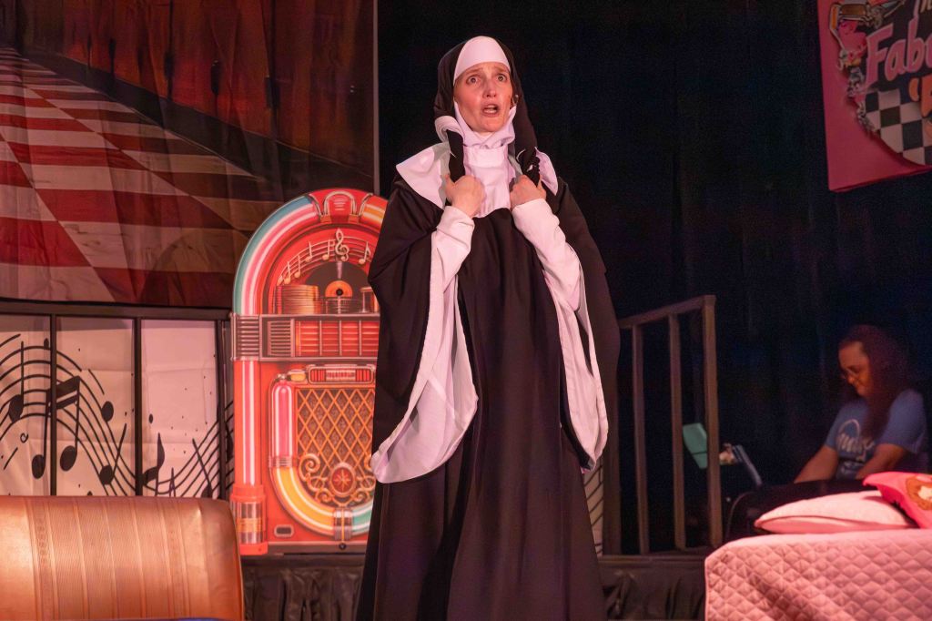 A woman dressed as a nun performs on stage, expressing emotion, with a jukebox and musical notes in the background.