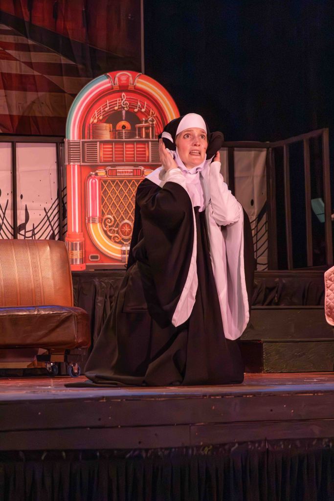 A performer dressed as a nun on stage, expressing distress while holding their ears, with a colorful vintage jukebox backdrop.