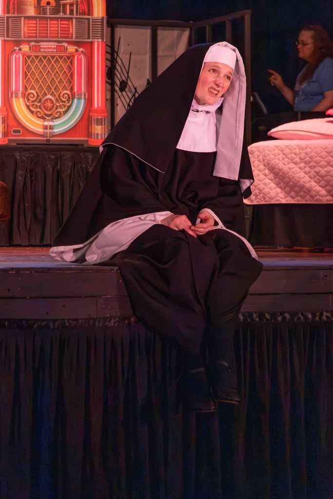 A performer dressed as a nun, sitting on a stage, with an expressive facial expression. Behind her is a jukebox and a partially visible bed.