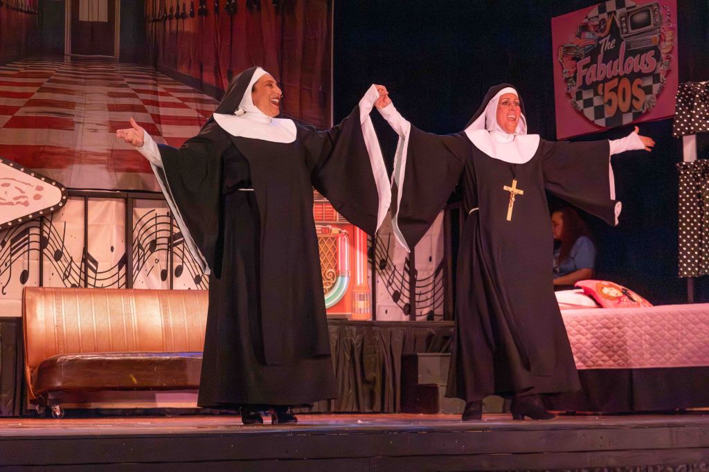 Two nuns in black and white habits joyfully performing on stage, holding hands and smiling, with a colorful backdrop featuring music notes and a vintage theme.