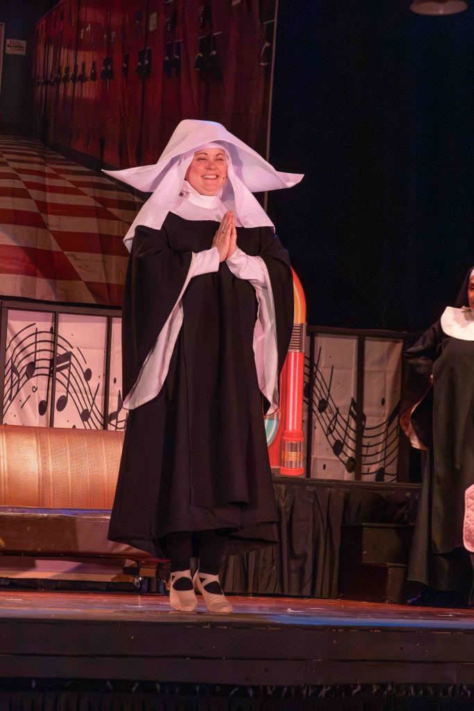 A performer dressed as a nun smiling and standing with hands in a prayer position on stage, with a colorful backdrop of lockers and musical notes.