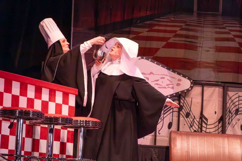 Two performers on stage, one dressed as a chef with a tall white hat and the other as a nun in a habit, with the chef holding a mixing bowl above the nun's head while she gestures with her hands. The background features a checkered bar and a musical motif.