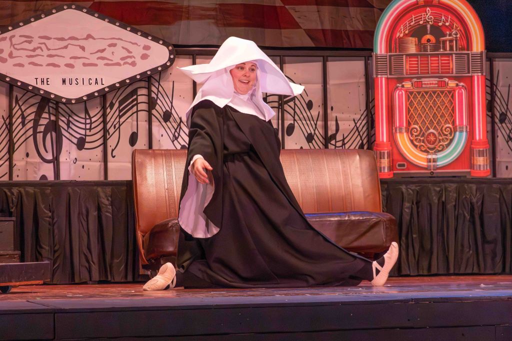 A performer in a black and white nun costume dancing on stage with a joyful expression, sitting on a brown couch with music-themed backdrop decorations.