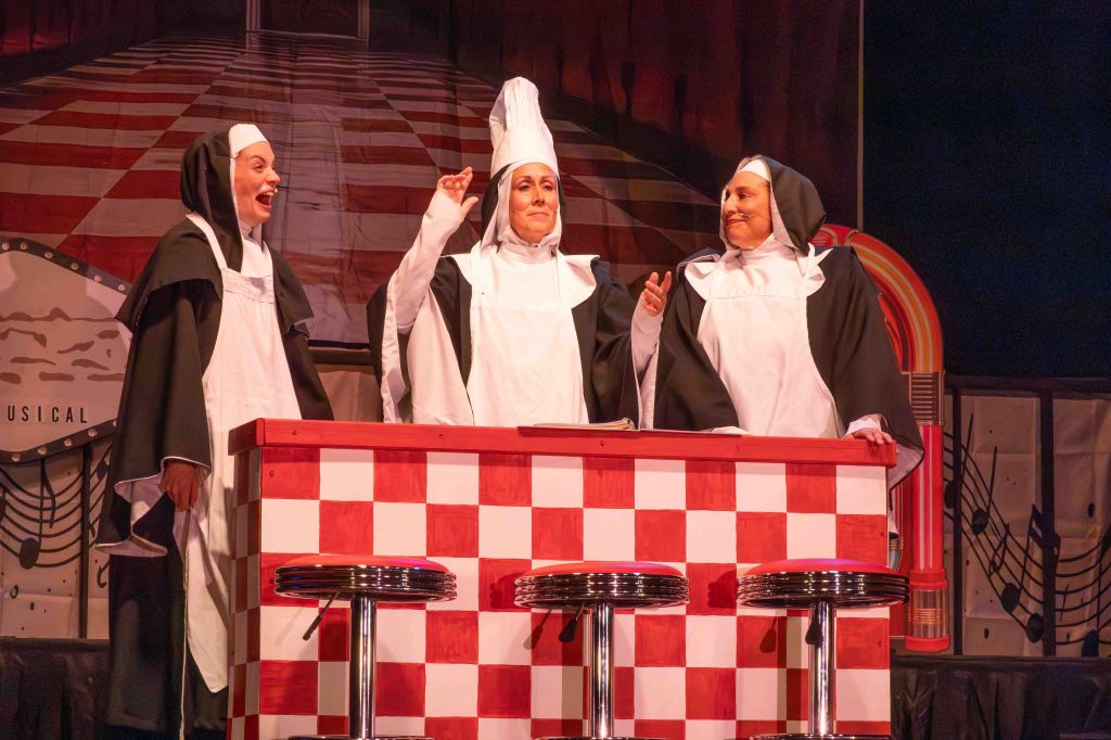 Three nuns in costumes performing on stage in front of a red and white checkered counter with bar stools.
