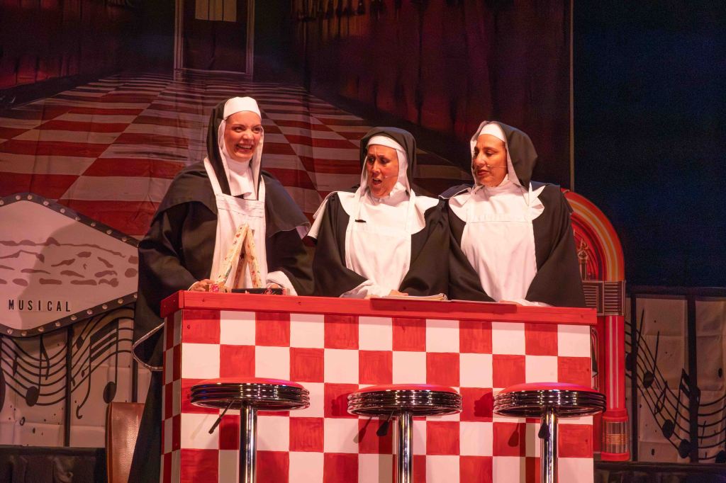 Three actresses dressed as nuns performing on stage at a musical, standing behind a red and white checkered counter with bar stools.
