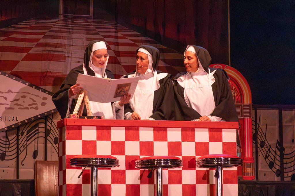 Three nuns in traditional attire stand around a checkered table, looking at a book, with a jukebox and musical-themed backdrop behind them.