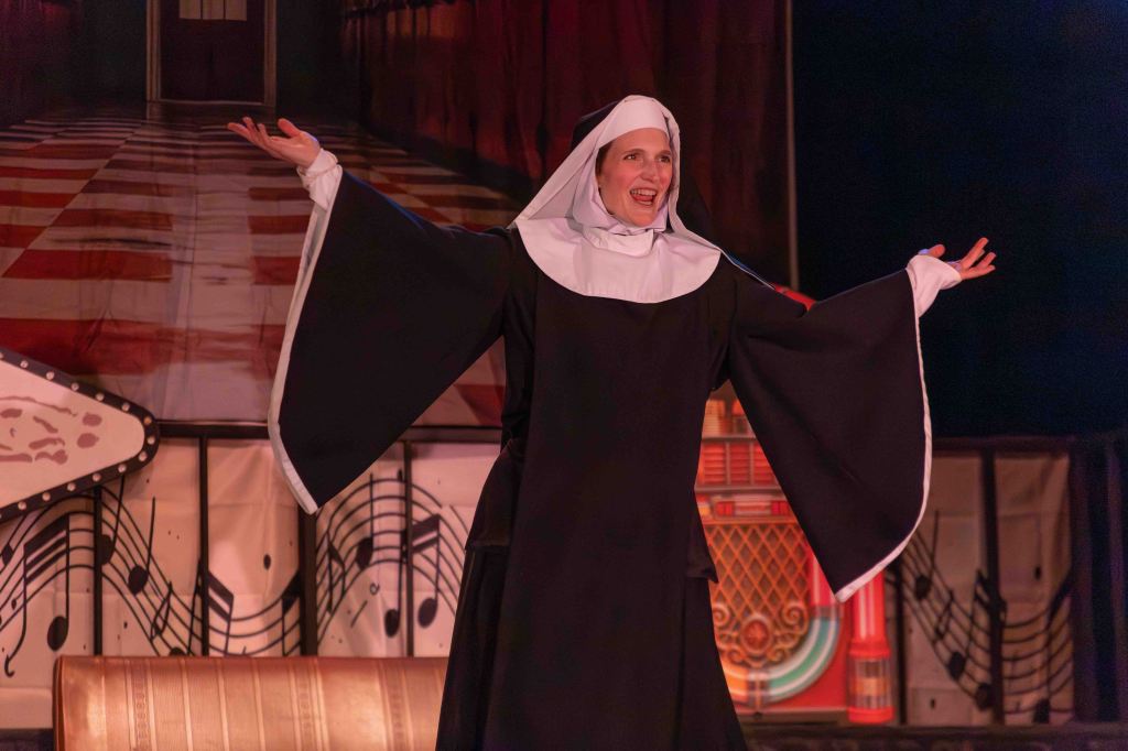 A performer dressed as a nun, smiling and gesturing with open arms on stage, with a colorful backdrop featuring musical notes and a vintage jukebox.