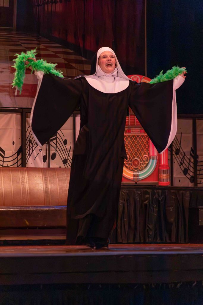 A performer dressed as a nun with outstretched arms, holding green feather boas, smiles joyfully on stage.