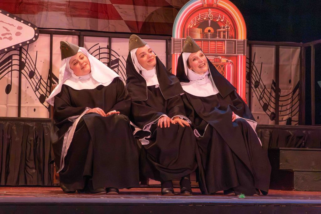 Three nuns in dark robes and white headdresses smiling and sitting on a stage decorated with musical notes and a jukebox.