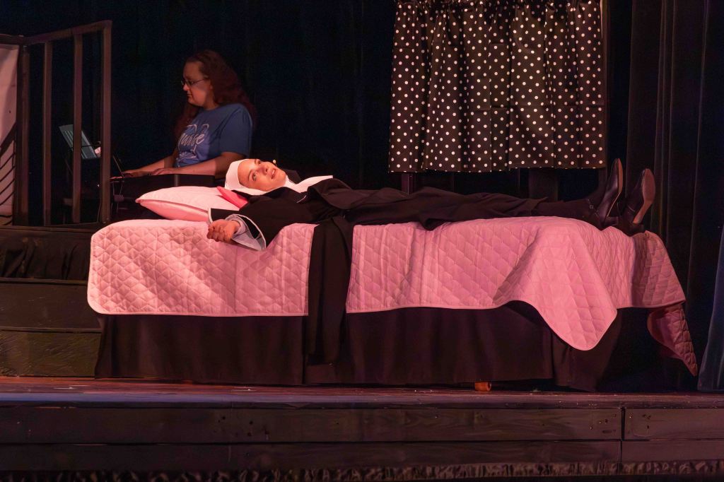 A scene from a theater production featuring a person in a nun's costume lying on a bed with a pink quilt, while another person in a blue shirt is seated nearby with a laptop.