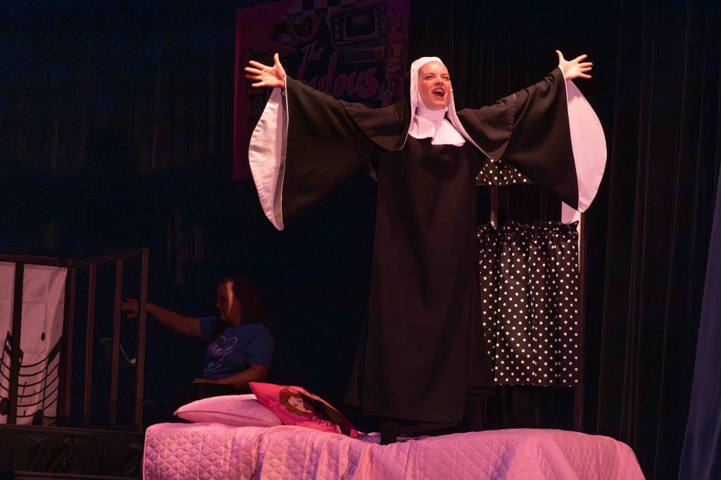A performer dressed as a nun dramatically poses on a bed, with arms outstretched, in a theatrical setting. A woman in a blue shirt is seen in the background.
