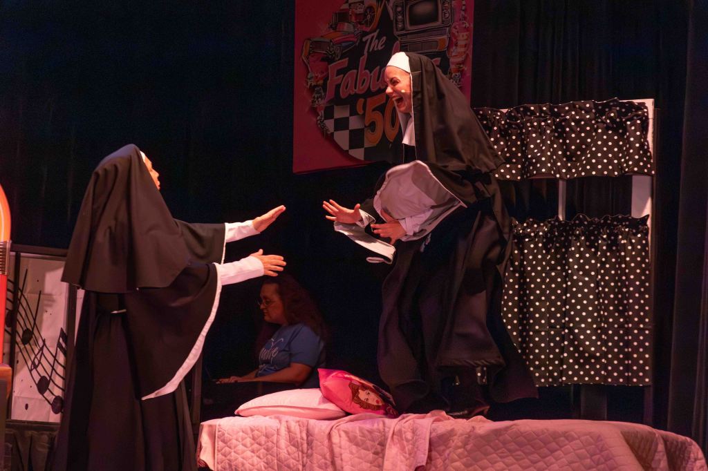 Two actresses dressed as nuns performing on stage, one reaching out to the other who is jumping excitedly, with a colorful backdrop and props in the scene.