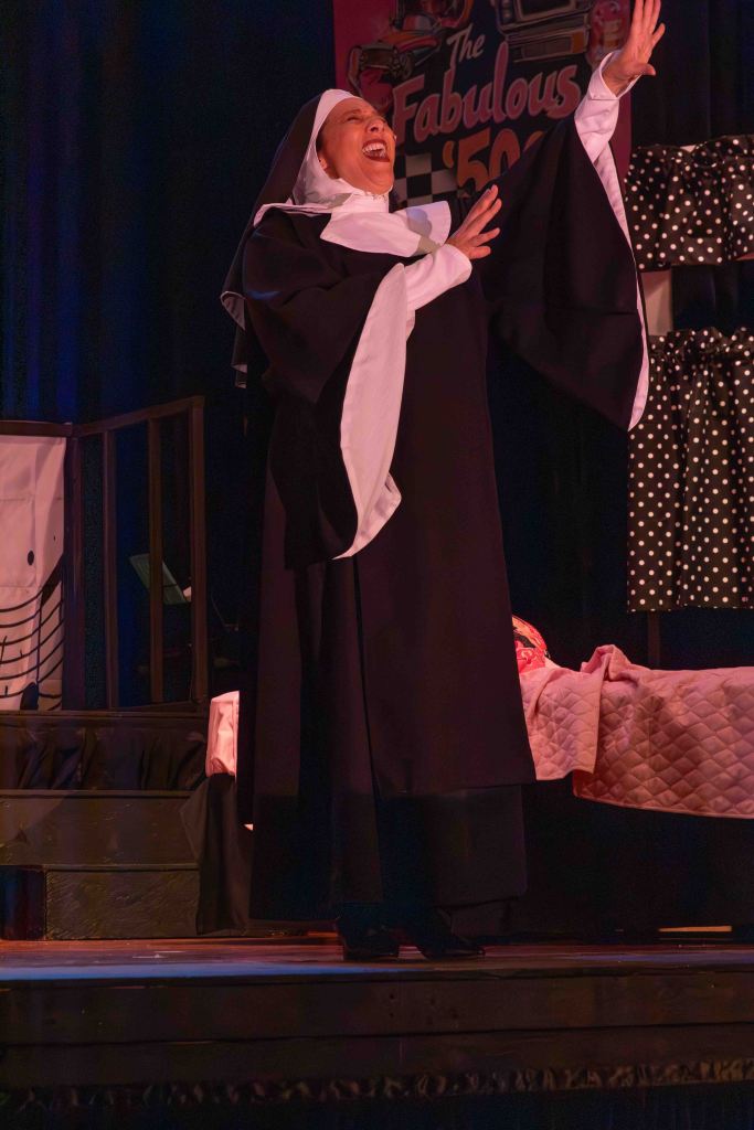 A performer dressed as a nun passionately singing on stage with a joyful expression, surrounded by a theater backdrop featuring a retro theme.