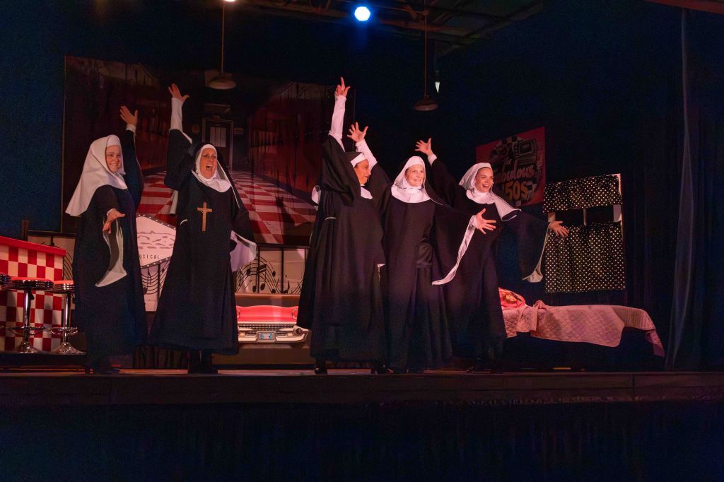 Five nuns in traditional black habits performing on stage, with a colorful backdrop featuring a retro diner and decorated set.