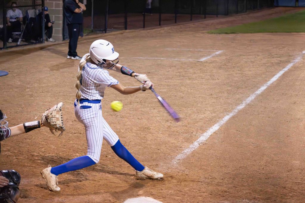 A young female softball player swings a bat to hit a yellow softball during a night game on a dirt field.