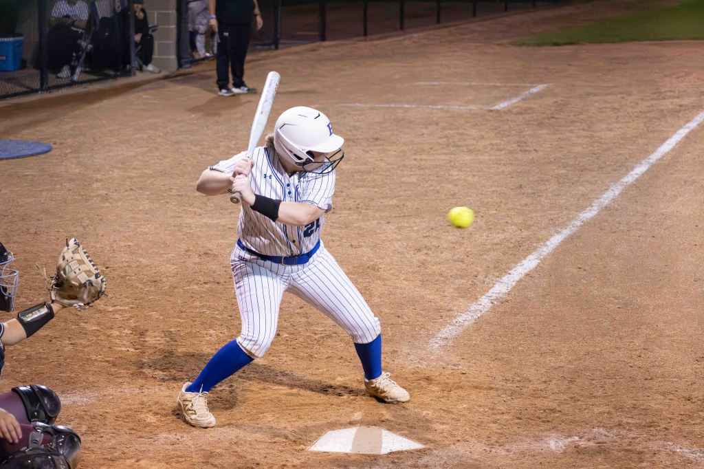 A female softball player swings at a pitch while standing at home plate on a dirt field.