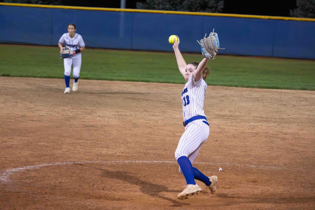 A softball player with the number 13 on her uniform is pitching the ball on a dirt field during a night game, while a teammate is in the background preparing to catch.