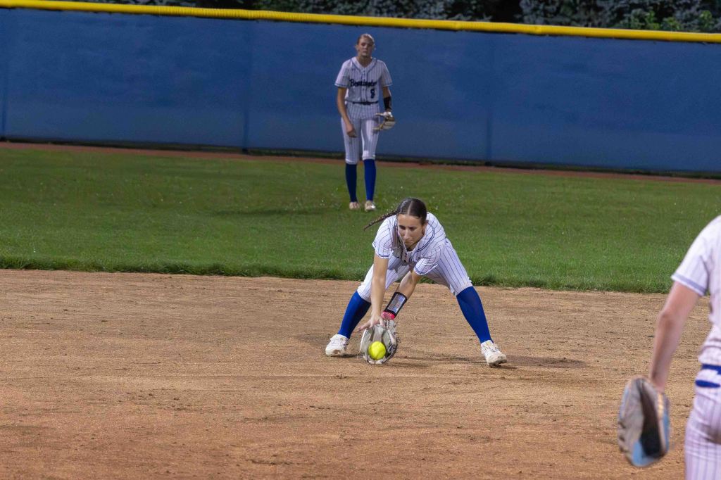 A female softball player crouching down to field a ball on the infield, wearing a striped uniform and blue socks, with another player in the background.
