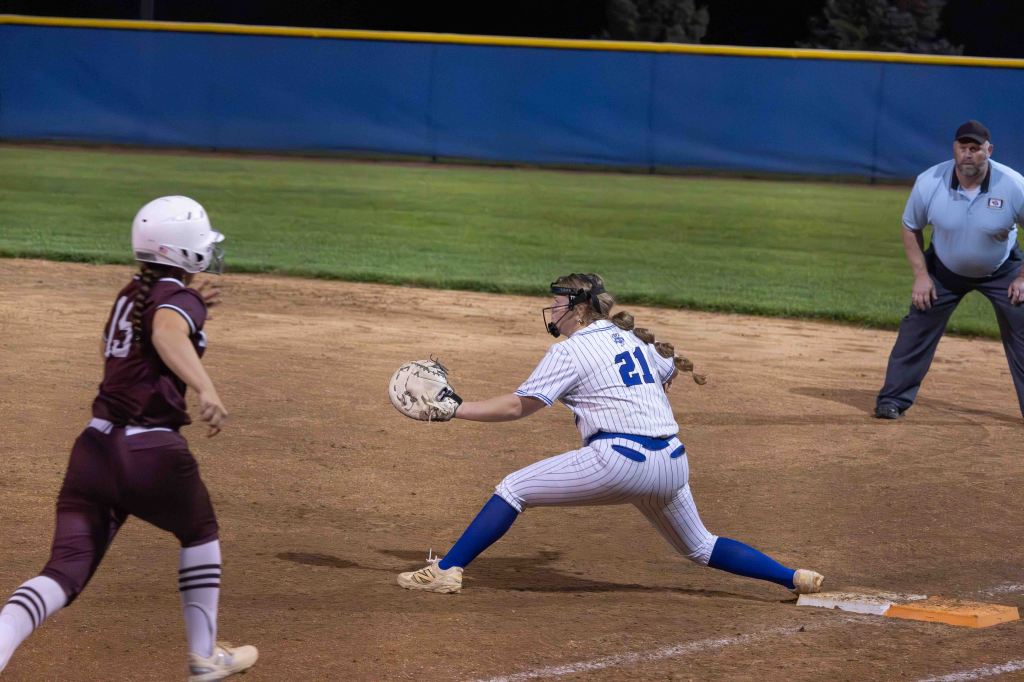 A female softball player in a blue and white striped uniform reaches for the ball at first base while another player in a maroon uniform runs towards her. An umpire observes the play in the background.
