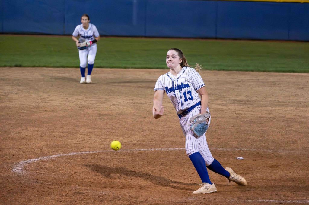 A softball player in a striped blue and white uniform throws a ball from the pitcher's mound while a teammate stands in the background.