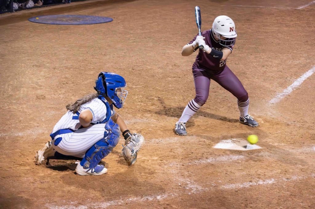 A baseball catcher in blue gear crouches behind home plate as a batter in maroon prepares to hit a pitch during a nighttime game.
