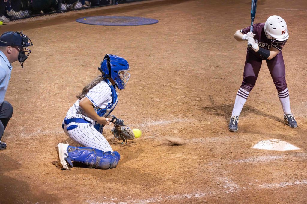 A softball game scene showing a catcher in blue gear preparing to catch a pitch while a batter in maroon is in the batting stance, with an umpire observing the play.
