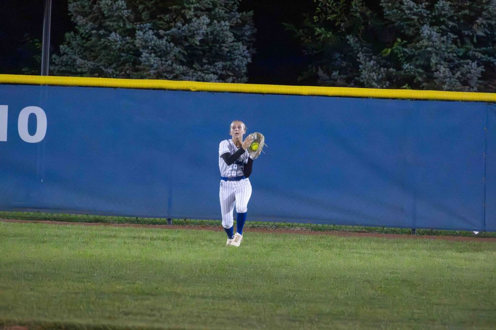 A softball player in a striped uniform stands in the outfield, preparing to catch a fly ball against a blue outfield wall.