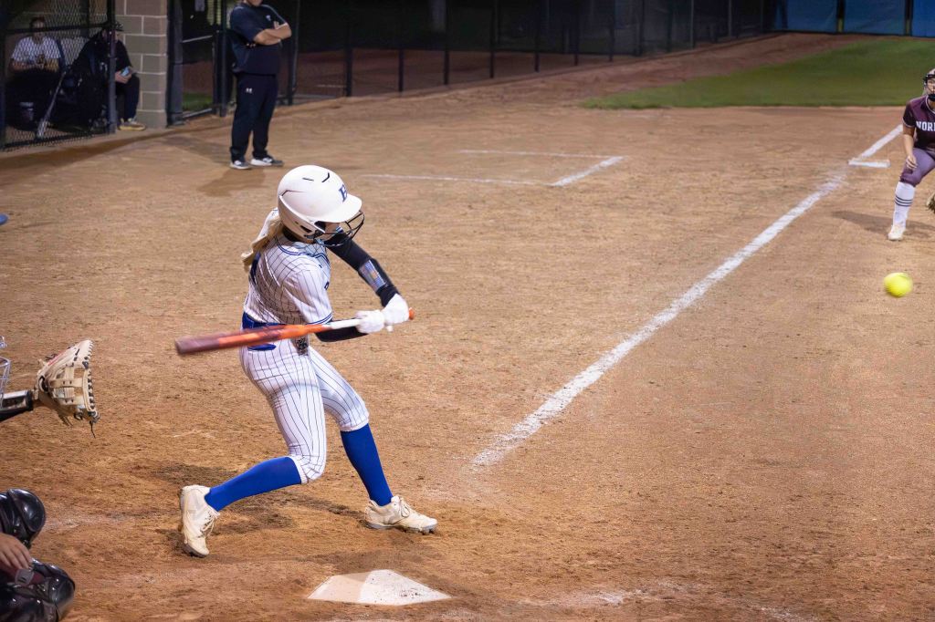 A female softball player in a white and blue uniform is swinging a bat to hit a yellow softball on a dirt field during a night game.