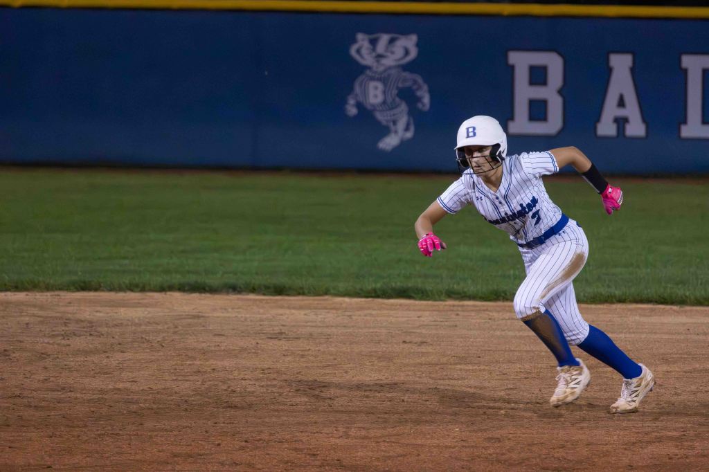 A female softball player in a striped uniform and helmet runs on the field during a game, with a focus on her athletic movement. The background features a team logo on a blue wall.