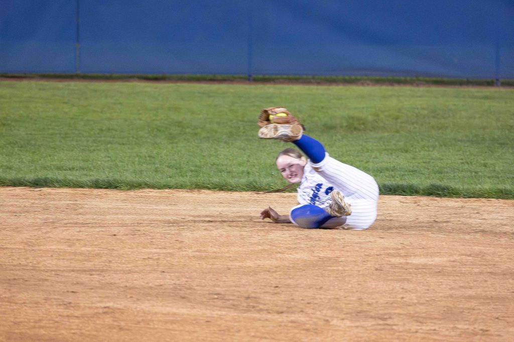 A softball player in a blue and white uniform is sliding on the ground while attempting to catch a ball, with a focused expression.