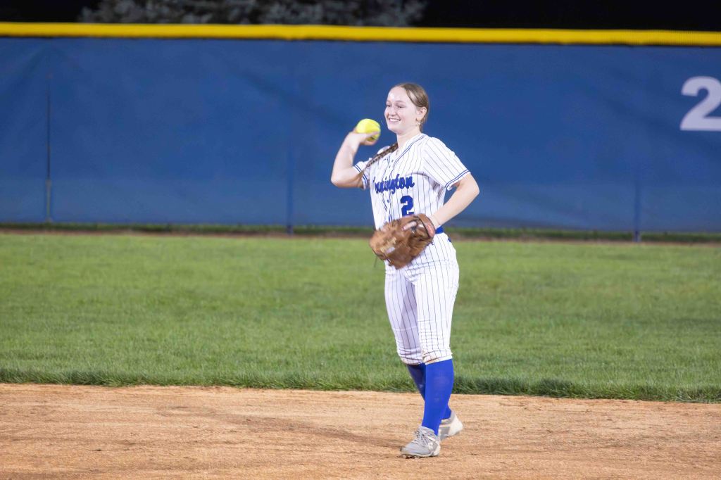 A female softball player in a striped white and blue uniform stands on the field, smiling while holding a softball and wearing a glove.