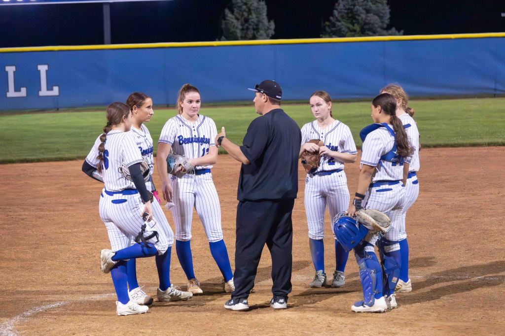 A softball coach discussing strategy with a group of players on the field during a night game.