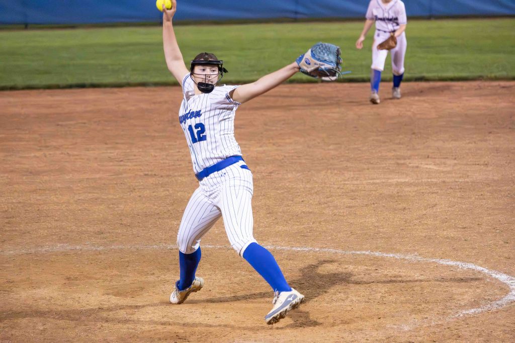 A female softball pitcher in a white and blue uniform winds up to throw a pitch, with one hand holding a yellow softball and the other in a throwing position.