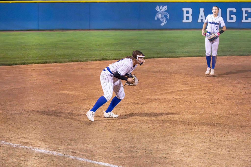 A female softball player in a crouched position, preparing to field a ball, with another player standing in the background. Both are wearing striped uniforms and blue socks.