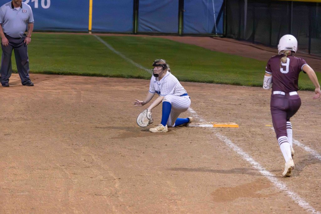 A softball game in action, showcasing a female player in a white uniform attempting to catch a ball while another player in maroon runs toward first base, with an umpire observing the play in the background.