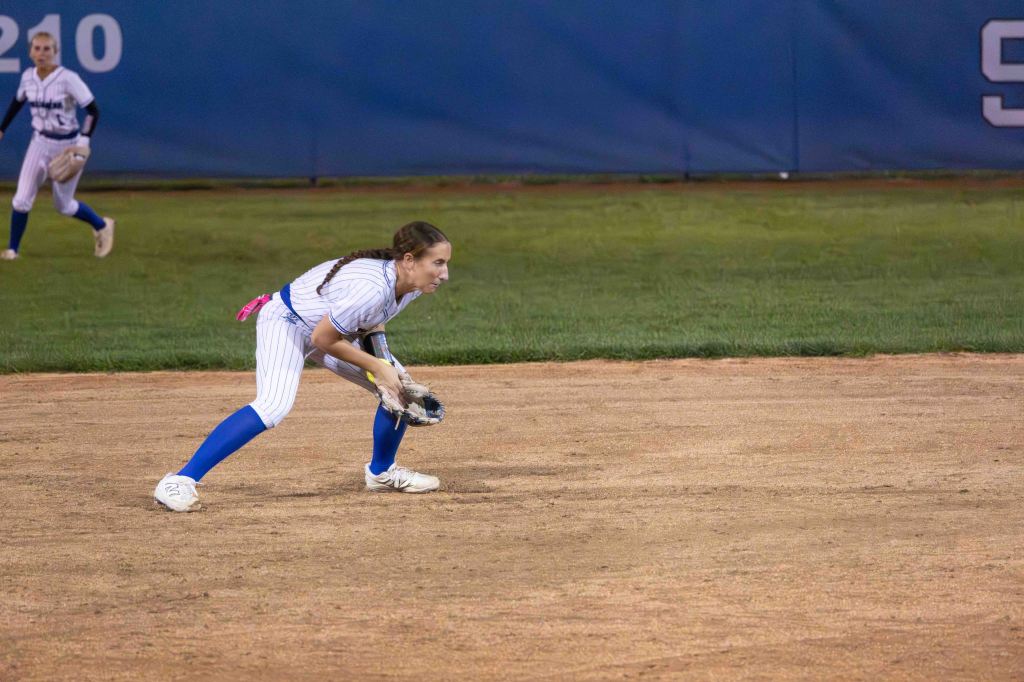 A female softball player crouched in a ready position on the infield dirt, wearing a white jersey with blue stripes and blue pants, focused on the play. Another player is visible in the background.