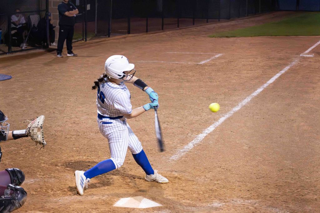 A female softball player with a bat is swinging at a yellow ball on a dirt field, with a catcher in the foreground and spectators in the background.